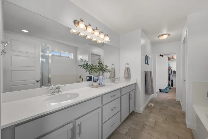 Dual vanity with integrated sinks, expansive mirror, wall-mounted lighting fixture, light-toned cabinetry, and large format floor tile