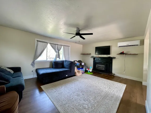Wood-finish flooring, ceiling fan, fireplace with dark surround, window with trim, and wall-mounted climate control unit