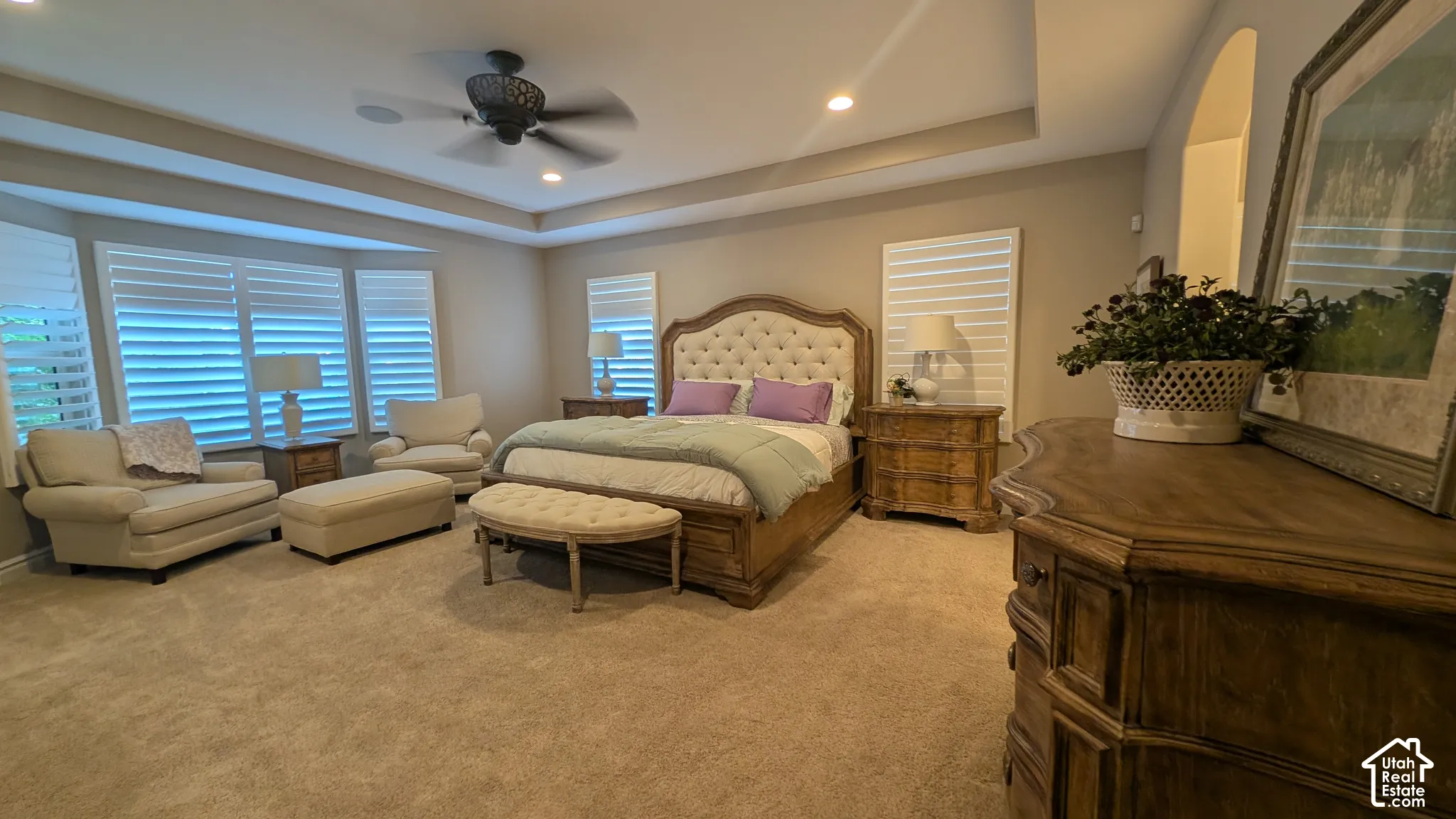Tray ceiling with recessed lighting, bay window with plantation shutters, ceiling fan, and carpeted flooring