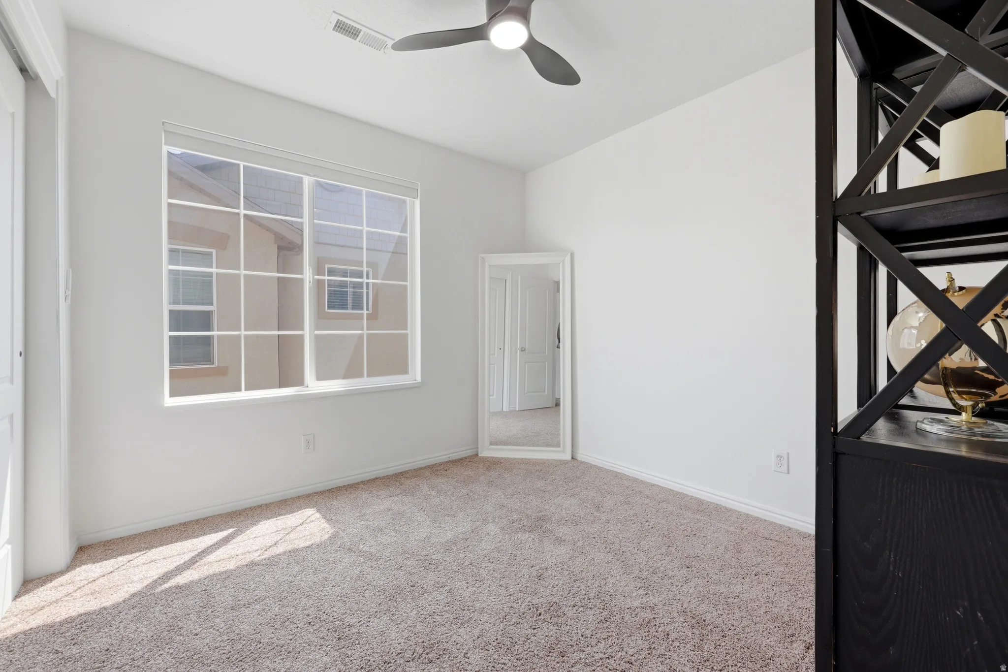 Carpeted flooring, ceiling fan with integrated lighting, large window with grid pattern, white interior walls, and built-in shelving unit