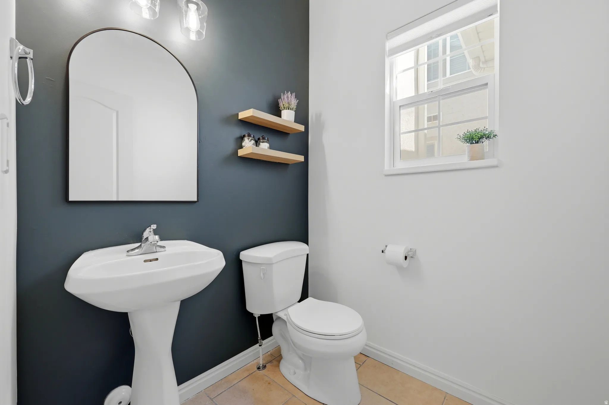 Pedestal sink, wall-mounted mirror with arched top, two floating wood shelves, and a single-pane window