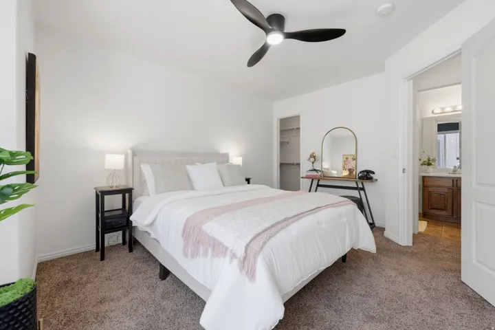 Primary bedroom featuring a vaulted ceiling with ceiling fan, carpeted flooring, dual nightstands with integrated lighting, and an ensuite bathroom with vanity and mirror visible through the adjacent doorway