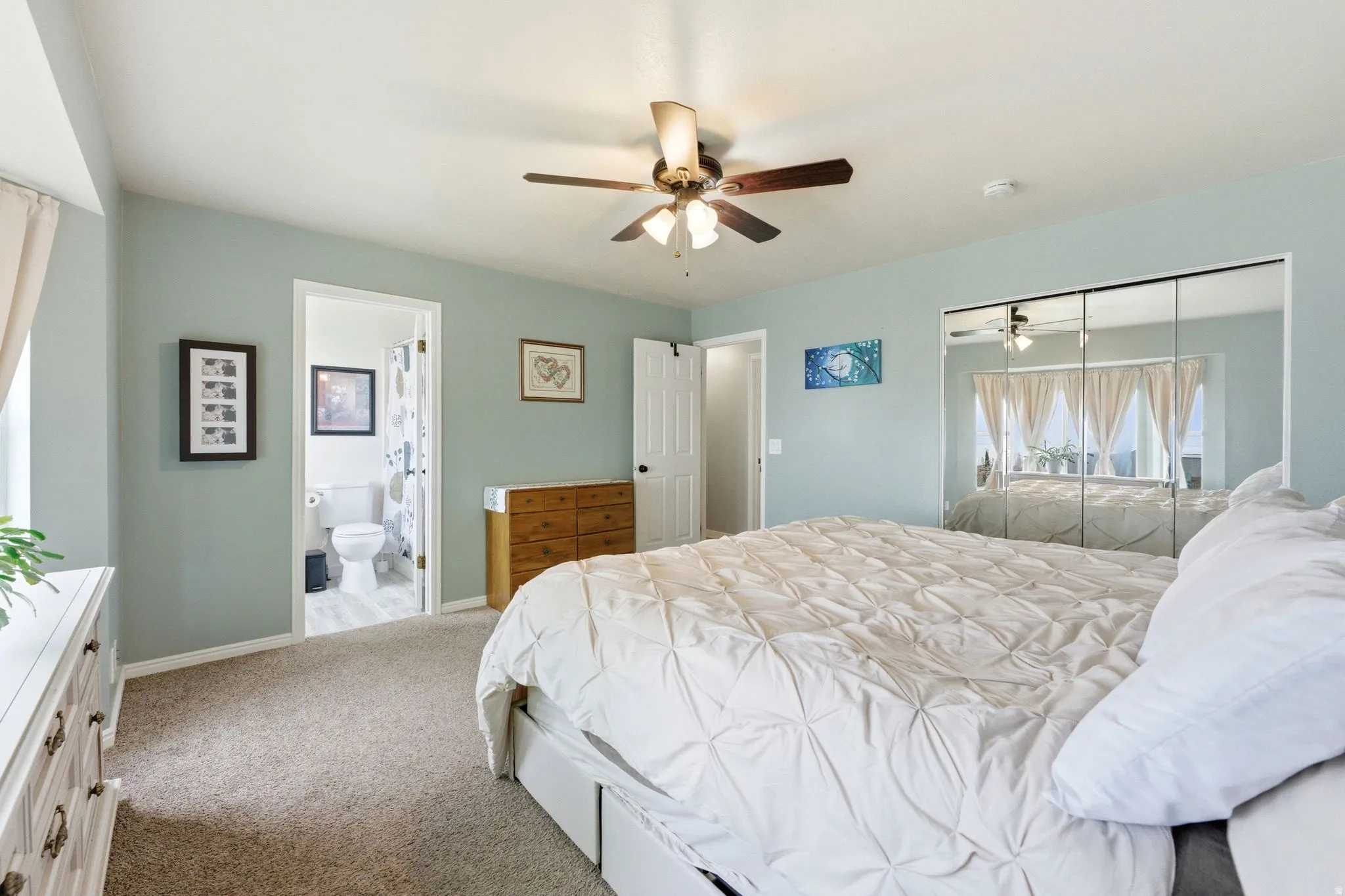 Carpeted bedroom featuring a ceiling fan, a mirrored closet, and an attached bathroom with hard-surface flooring