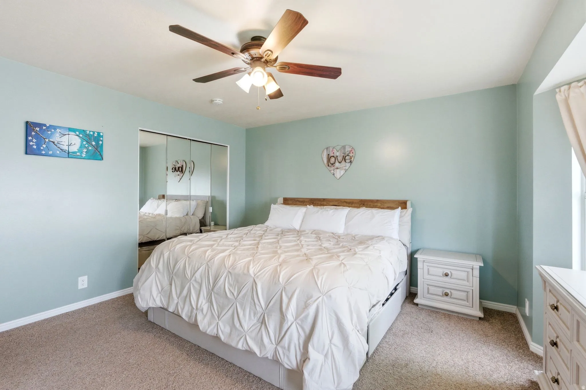 Ceiling fan with light fixture, mirrored closet doors, wood-finish headboard, window with curtain rod, and light-colored carpeting