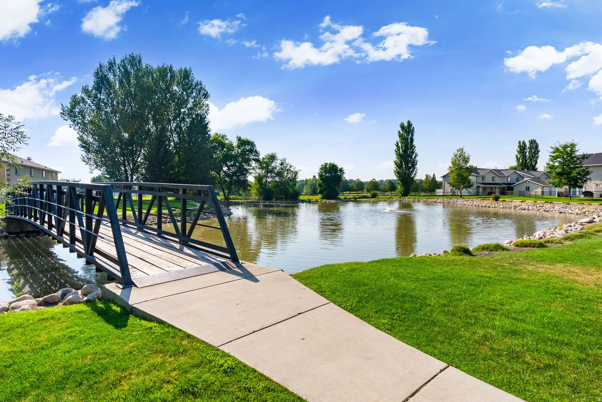 Pond with fountain feature, surrounded by green landscaping, a pedestrian bridge with metal railings and wood-finish decking, and residential structures in the background