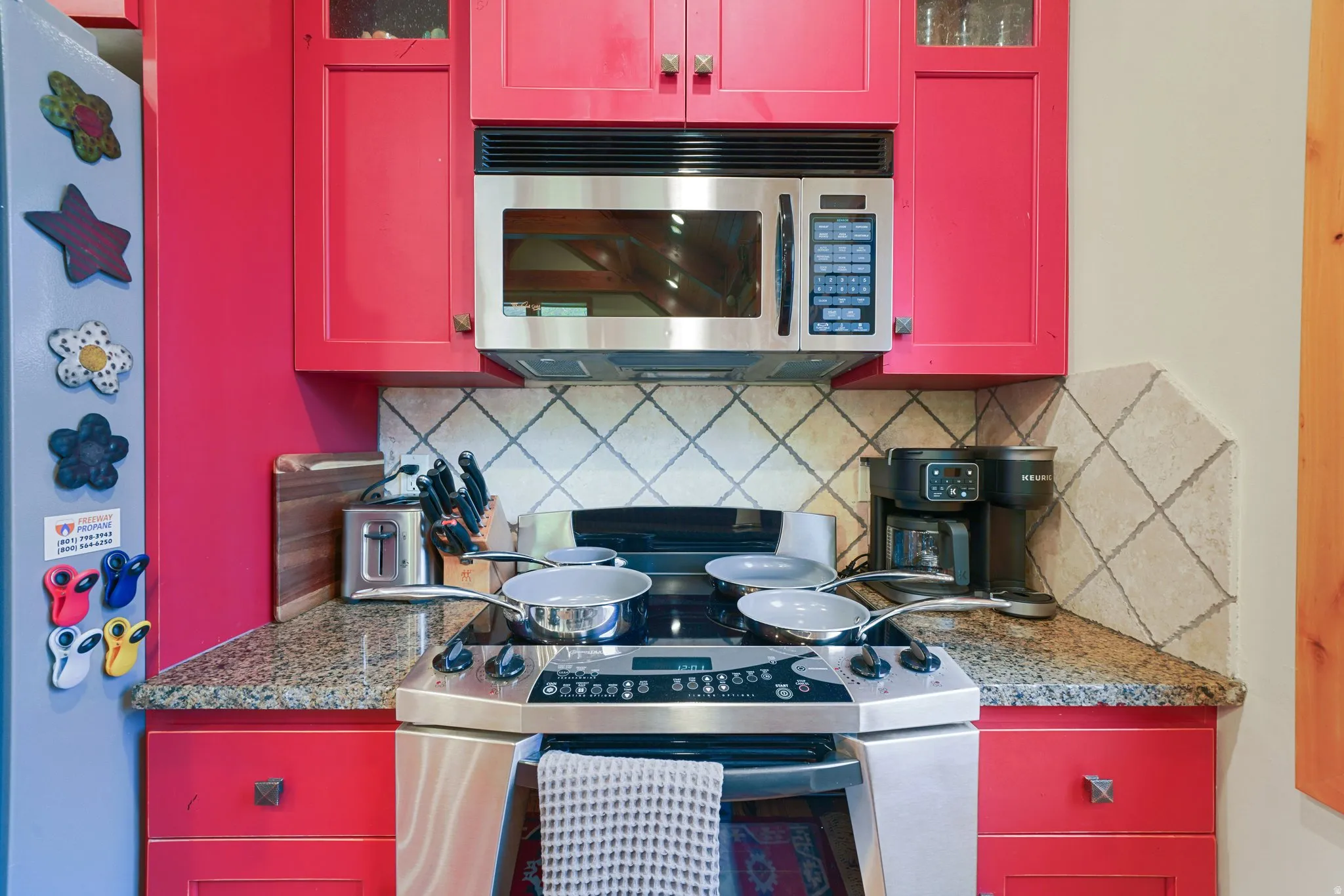 Red cabinetry with stainless steel hardware, granite-style countertops, stainless steel microwave, and a tile backsplash