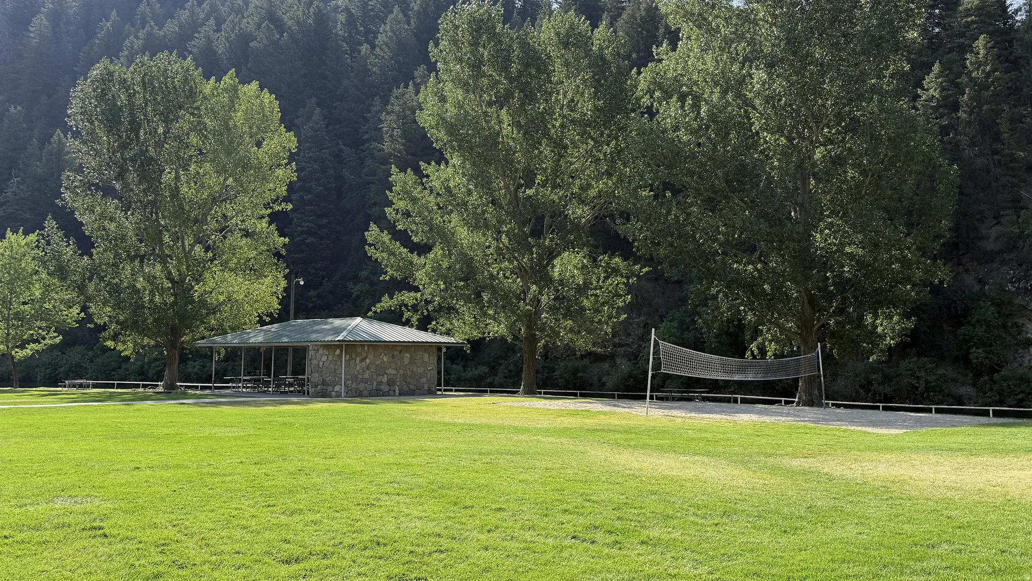 Expansive grassy area with mature trees, stone-sided picnic shelter, and a sand volleyball court