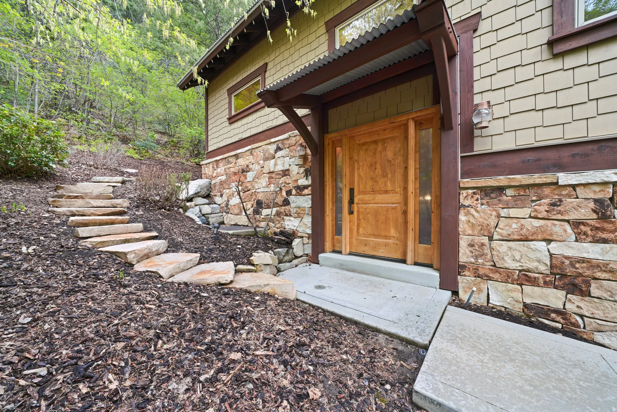 Natural wood entry door with vertical sidelights, stone veneer facade, shingle siding, and a corrugated metal awning