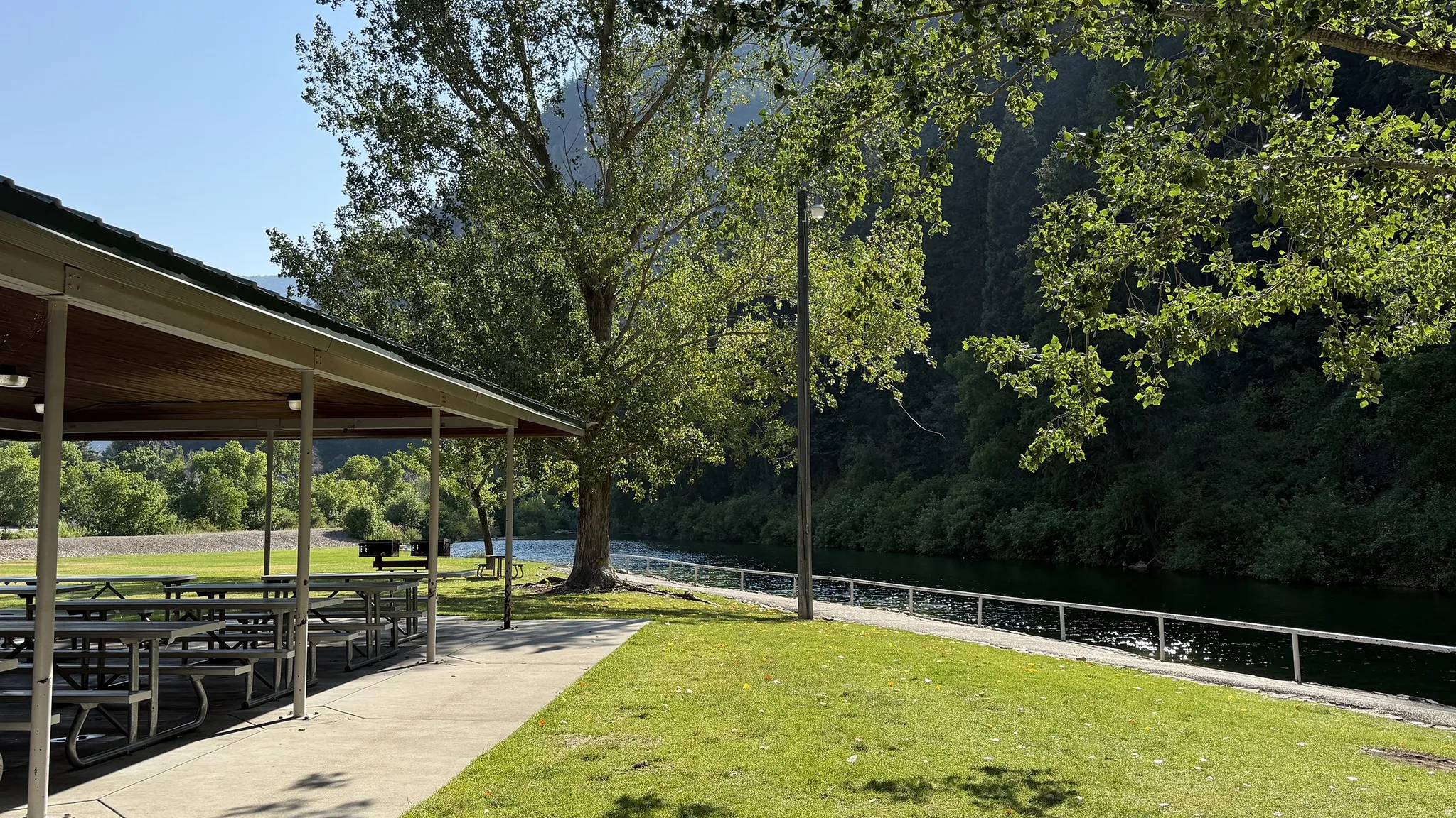 Covered outdoor recreational area with multiple picnic tables, a concrete walkway, and expansive green space bordering a body of water
