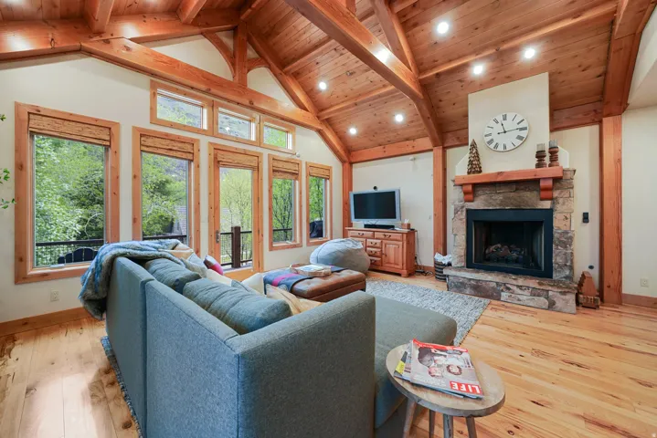 Vaulted wood-paneled ceiling with exposed beams, wood-finish flooring, stone-faced fireplace with wood mantel, multiple windows with wood trim, and recessed lighting