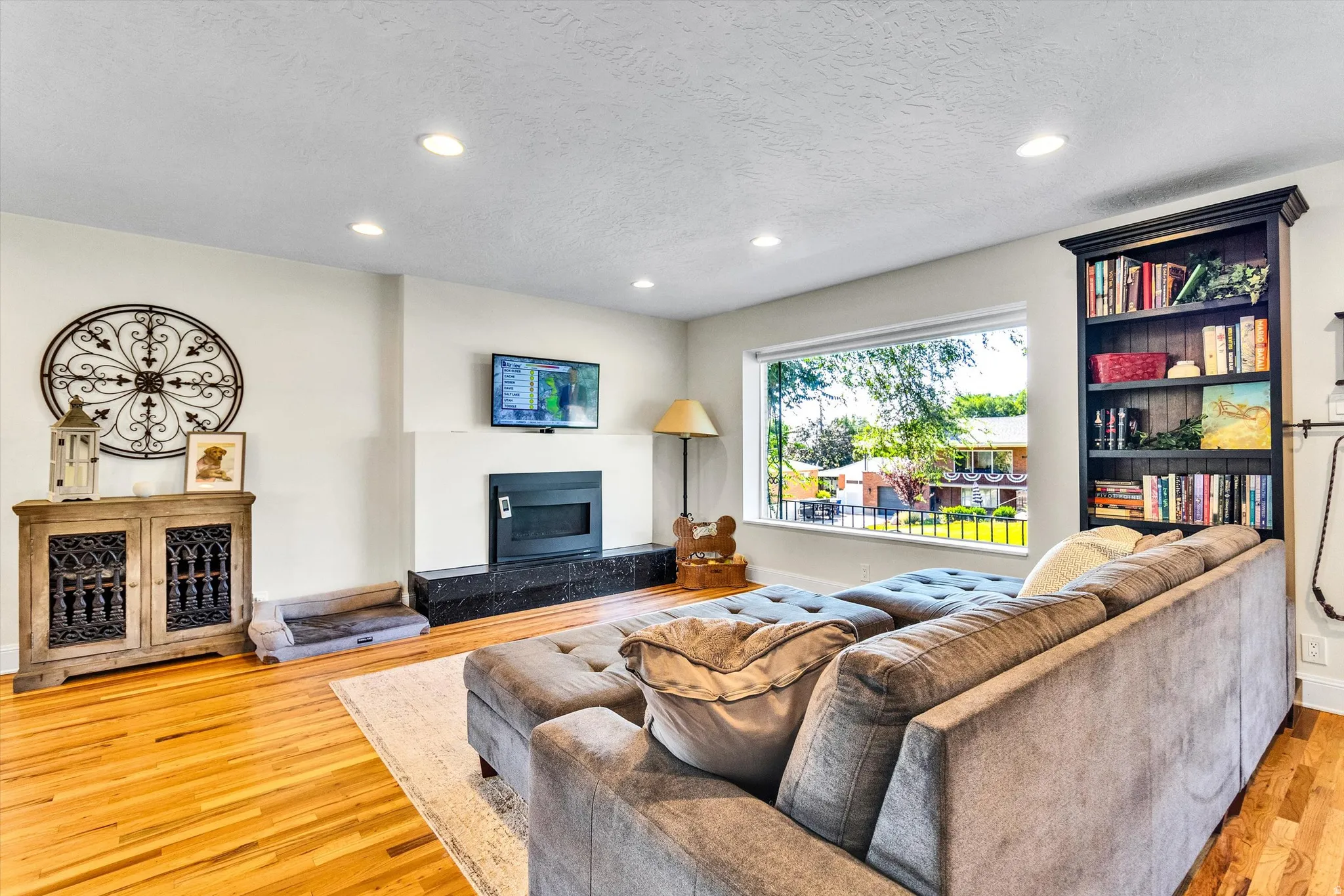 Living area featuring hardwood flooring, a fireplace with a black stone hearth, recessed lighting, a built-in bookshelf, and a large picture window