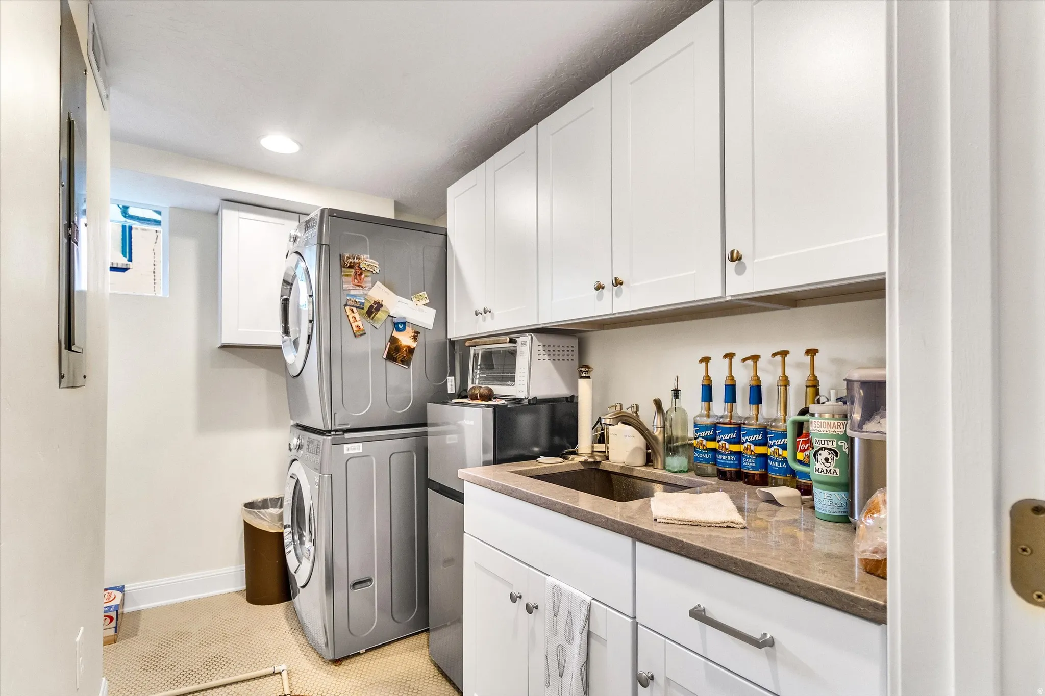 Laundry area featuring a stacked washer and dryer, white cabinetry with brass-toned hardware, a built-in sink, and a light-toned countertop