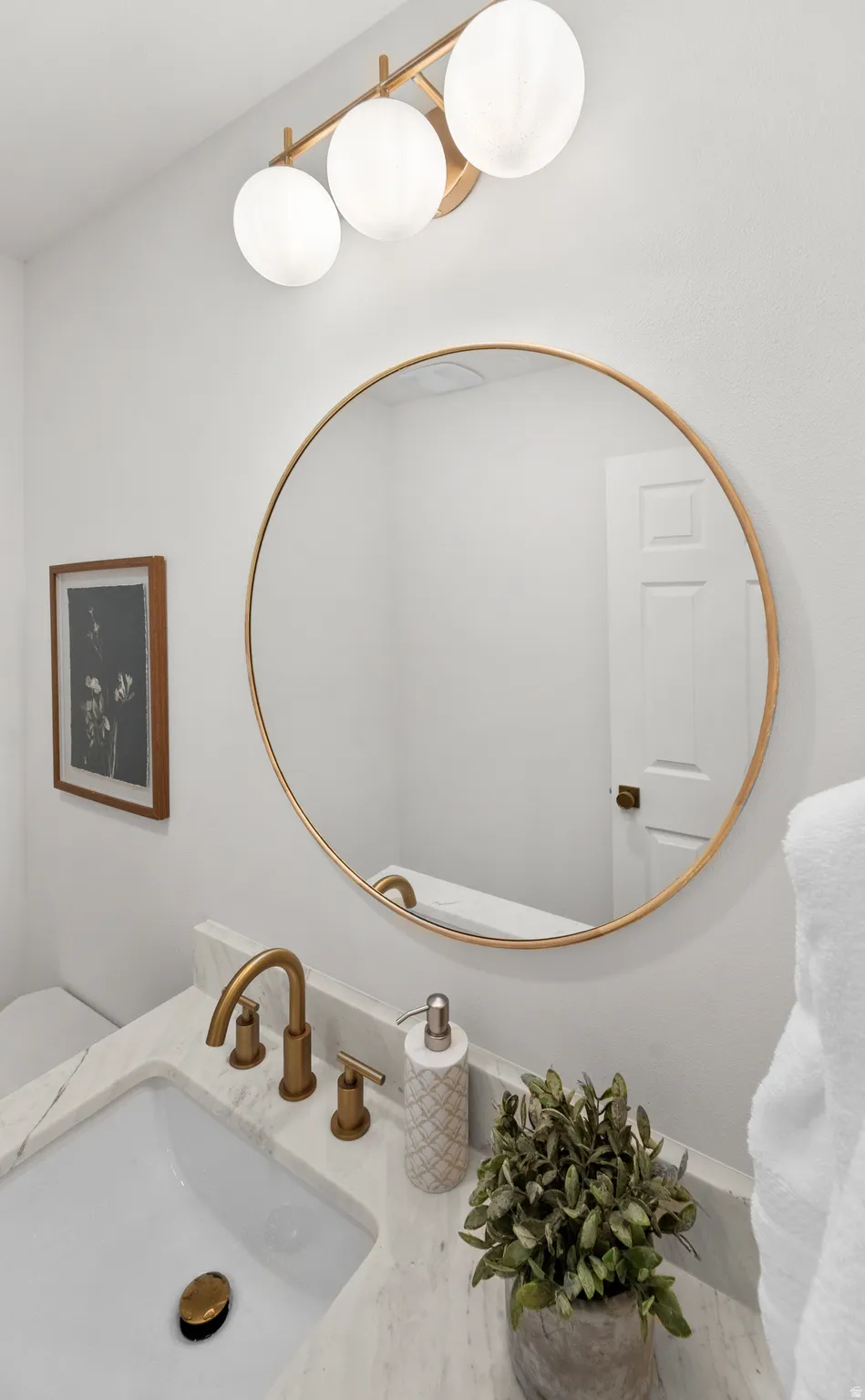 Gold-finish circular mirror, modern three-light vanity fixture, white wall-mounted toilet, gold-finish faucet, and marble-finish countertop