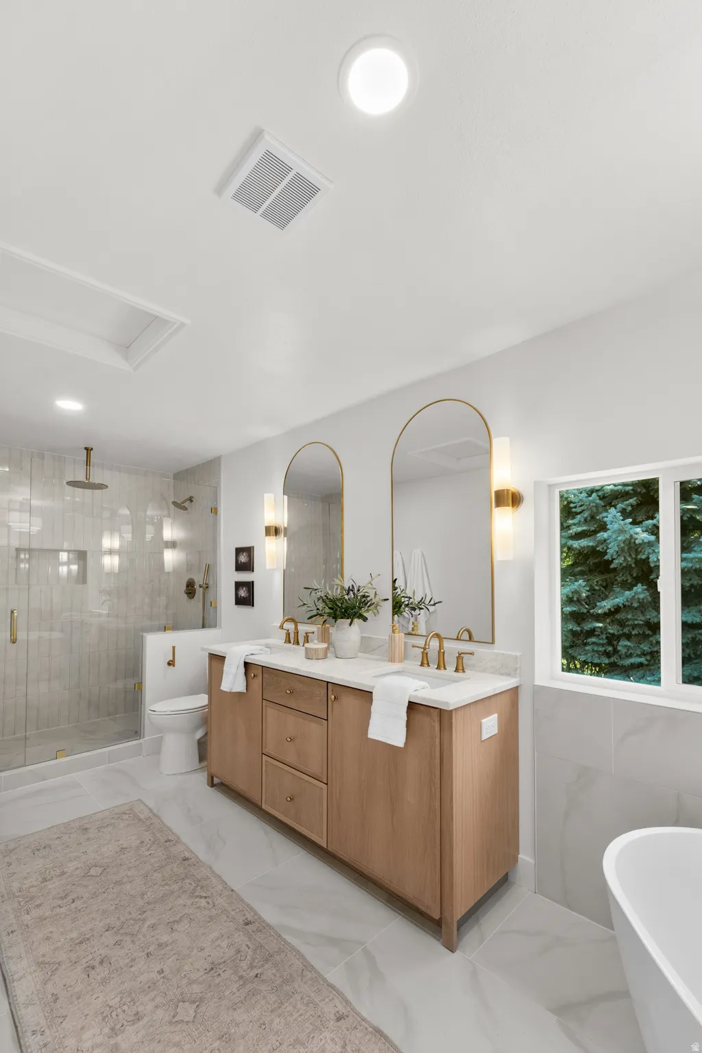 Dual-sink vanity with wood-finish cabinetry, a freestanding soaking tub, a walk-in shower with a glass enclosure, and large format floor tiles