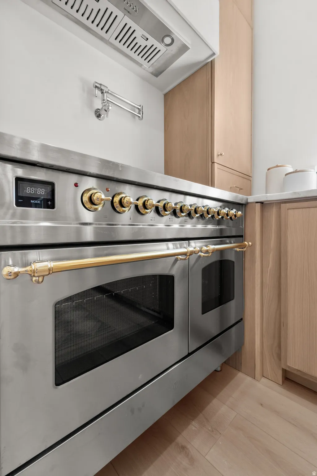 Stainless steel range with brass-finish hardware, stainless steel range hood, wall-mounted pot filler, light wood-finish cabinetry, and light wood-finish flooring