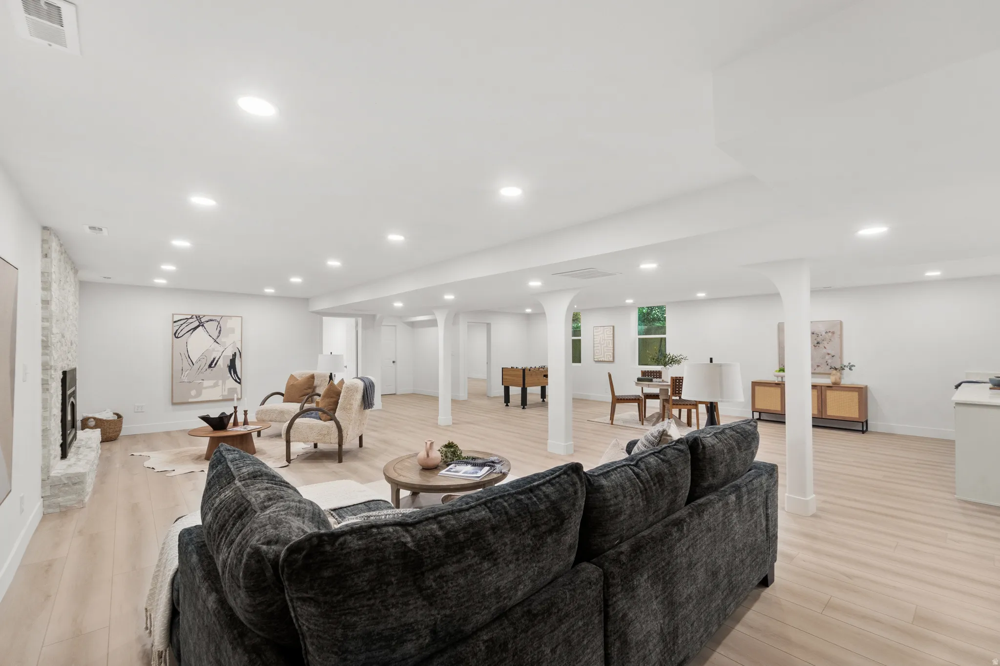 Open-concept living area with light wood-finish flooring, recessed lighting, white walls, and a stone-clad fireplace