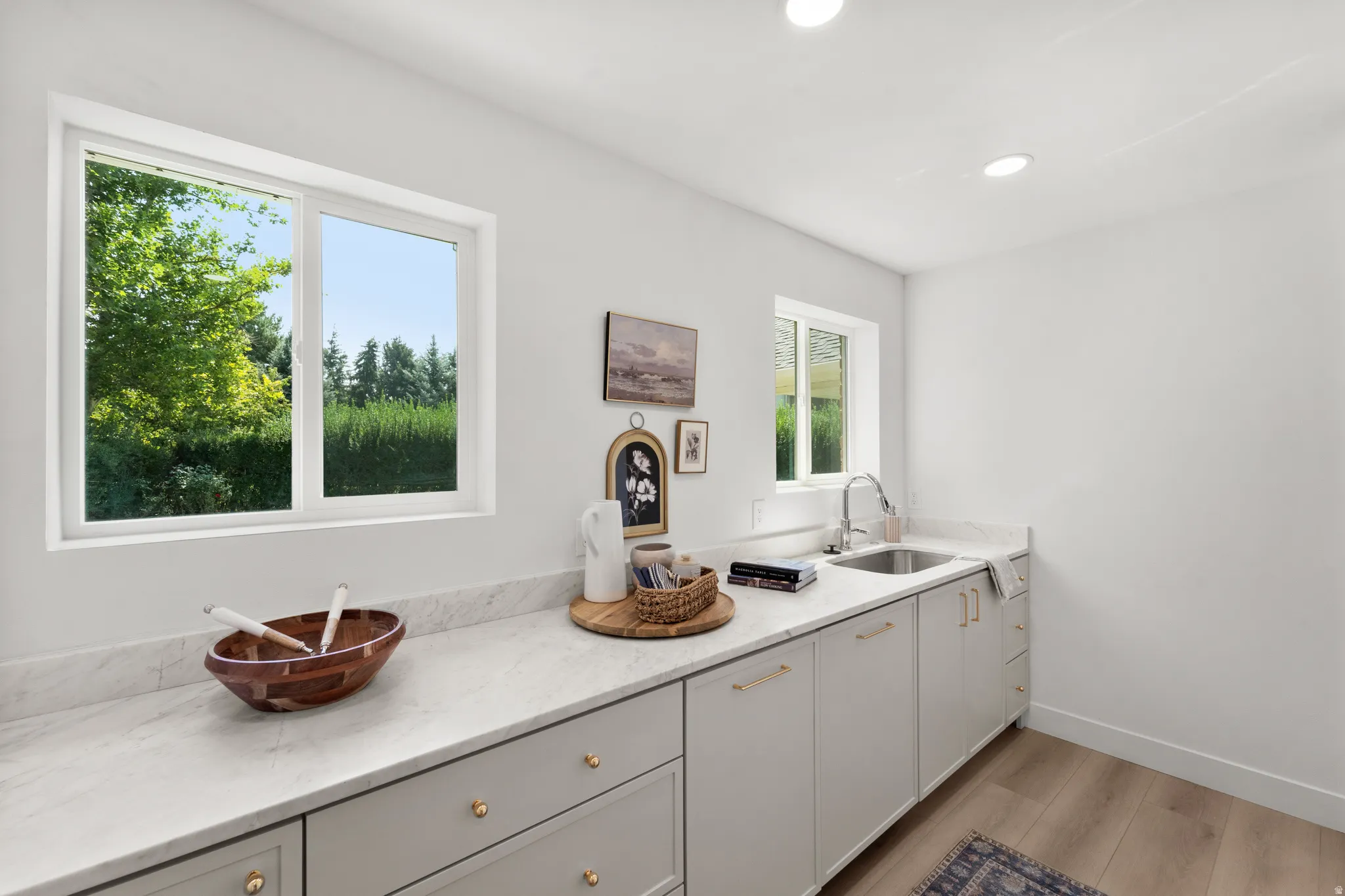 White walls with recessed lighting, light wood-finish flooring, white-framed windows, light-toned countertops, and gray cabinetry with gold-toned hardware