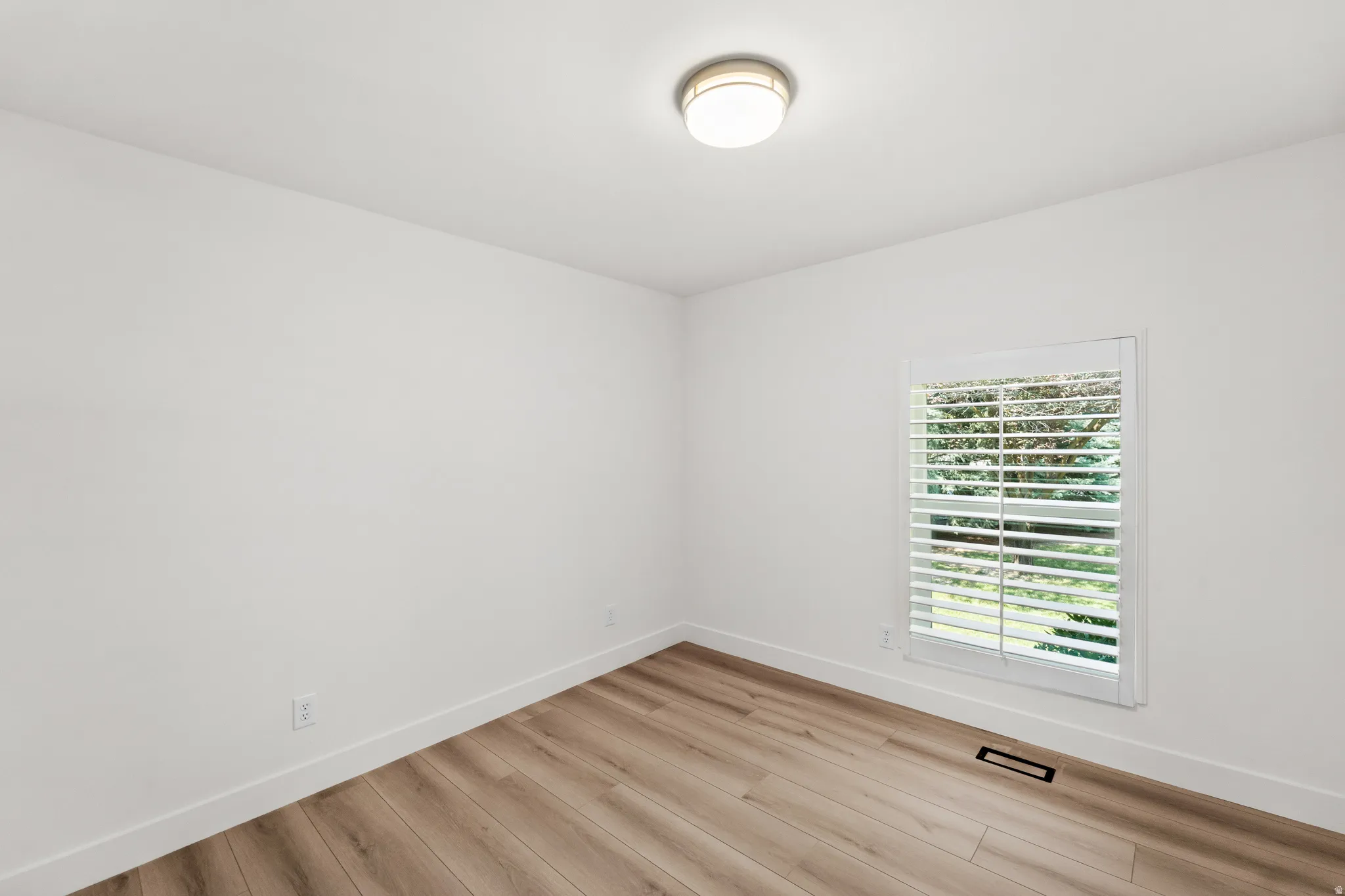 White interior walls with wood-finish flooring, a flush-mount ceiling light, and a window with white louvered shutters