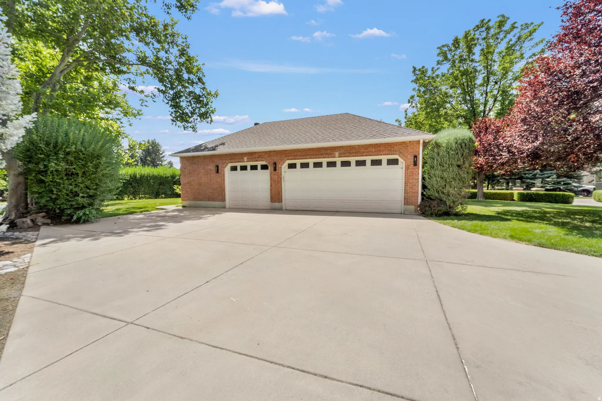 Brick exterior garage with two white garage doors, a concrete driveway, and surrounding mature landscaping
