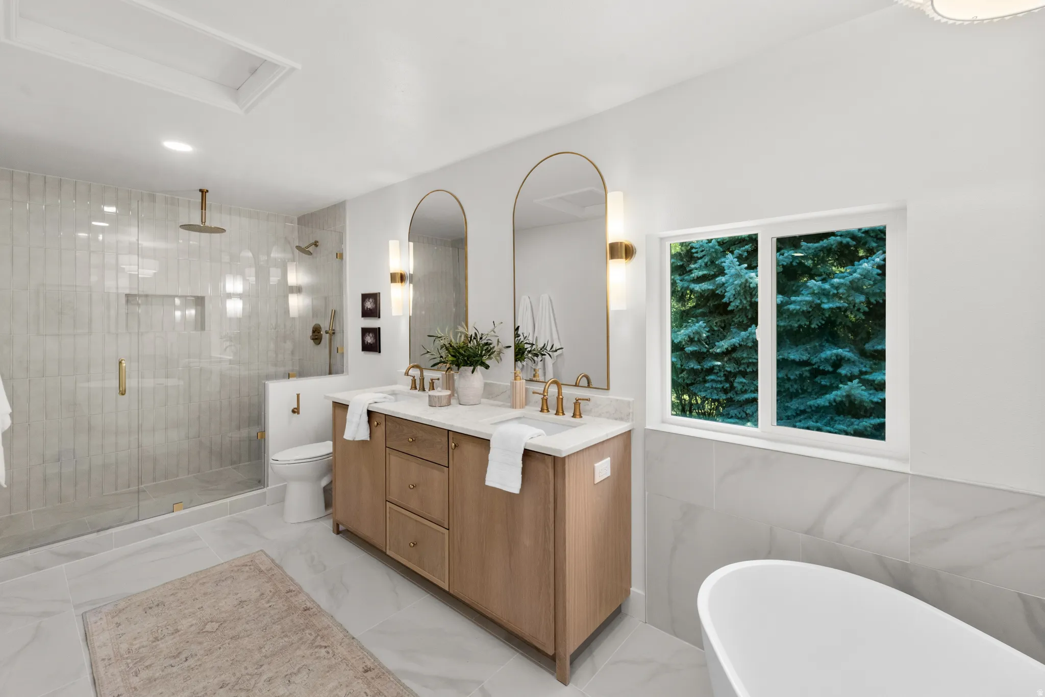 Spa-inspired primary bathroom featuring dual arched mirrors with brass fixtures, a wood-finish vanity with white quartz countertop, and polished concrete flooring