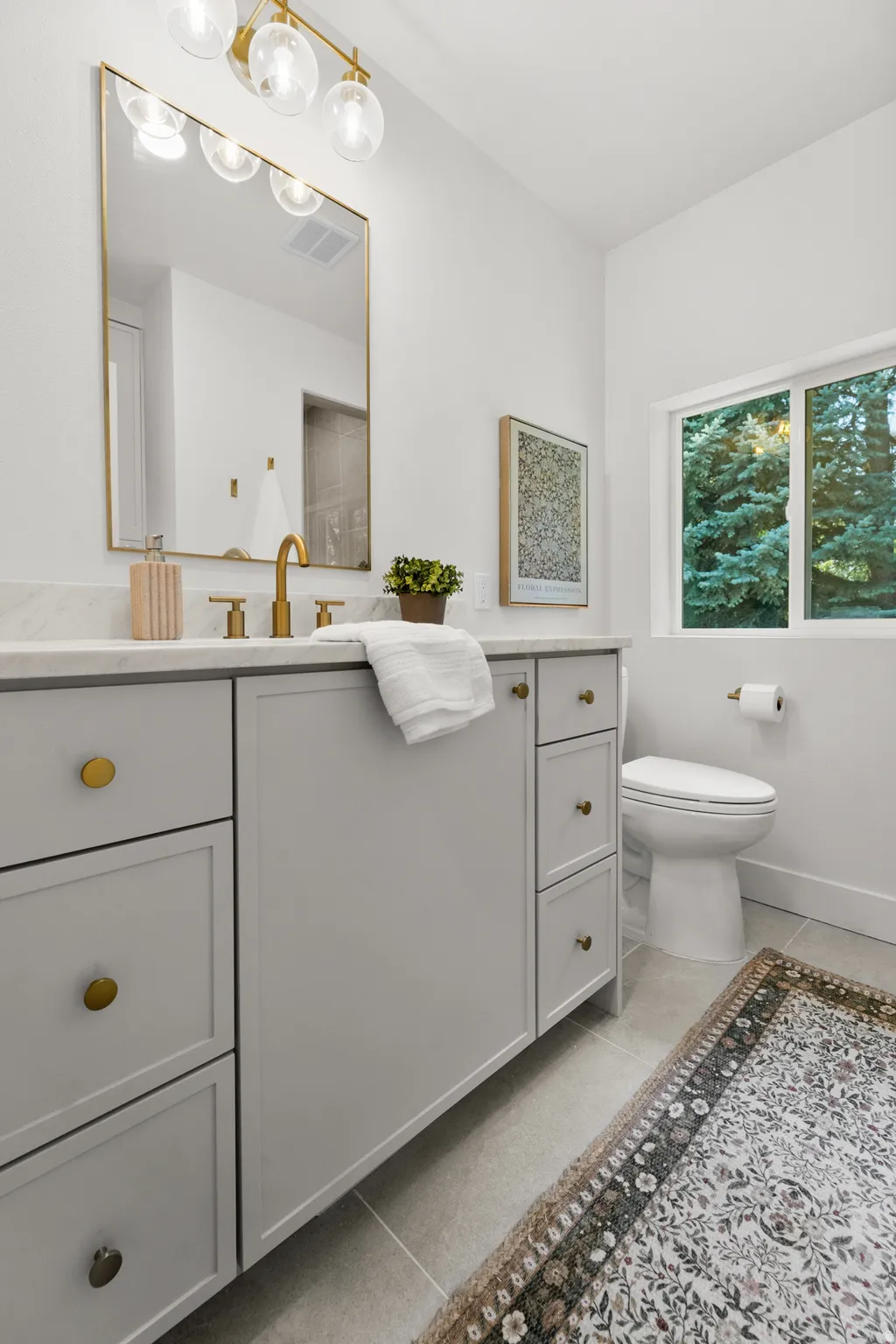 Light gray vanity with gold hardware, wide rectangular mirror with gold frame, gold-finish faucet, and window with natural scenery