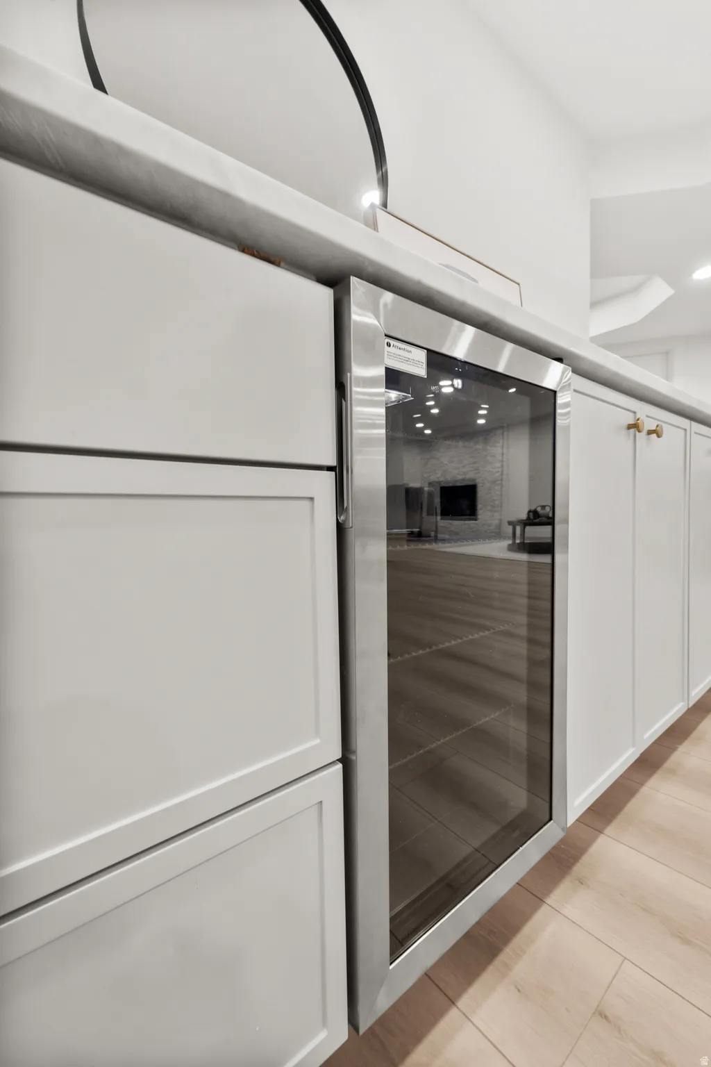 Built-in stainless steel beverage cooler, light-toned wood-finish flooring, white flat-panel cabinetry with gold-toned hardware, and a light-toned countertop