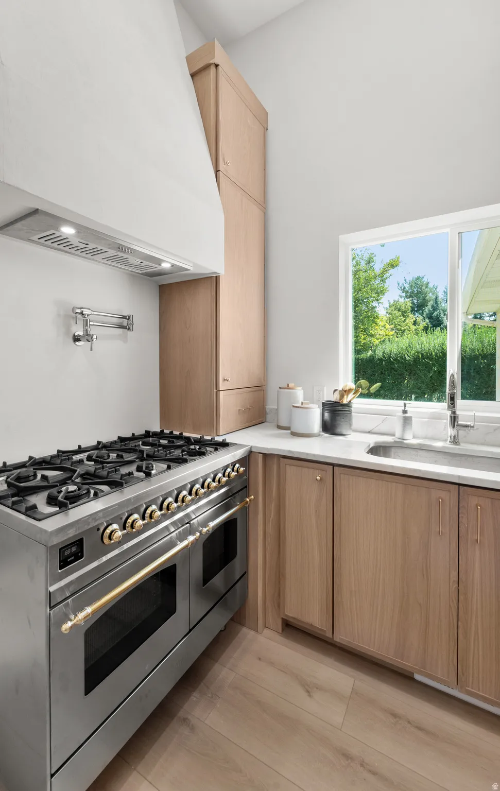 Stainless steel range with gold-finish hardware, pot filler, wood-finish cabinetry, light-colored countertops, and wood-finish flooring