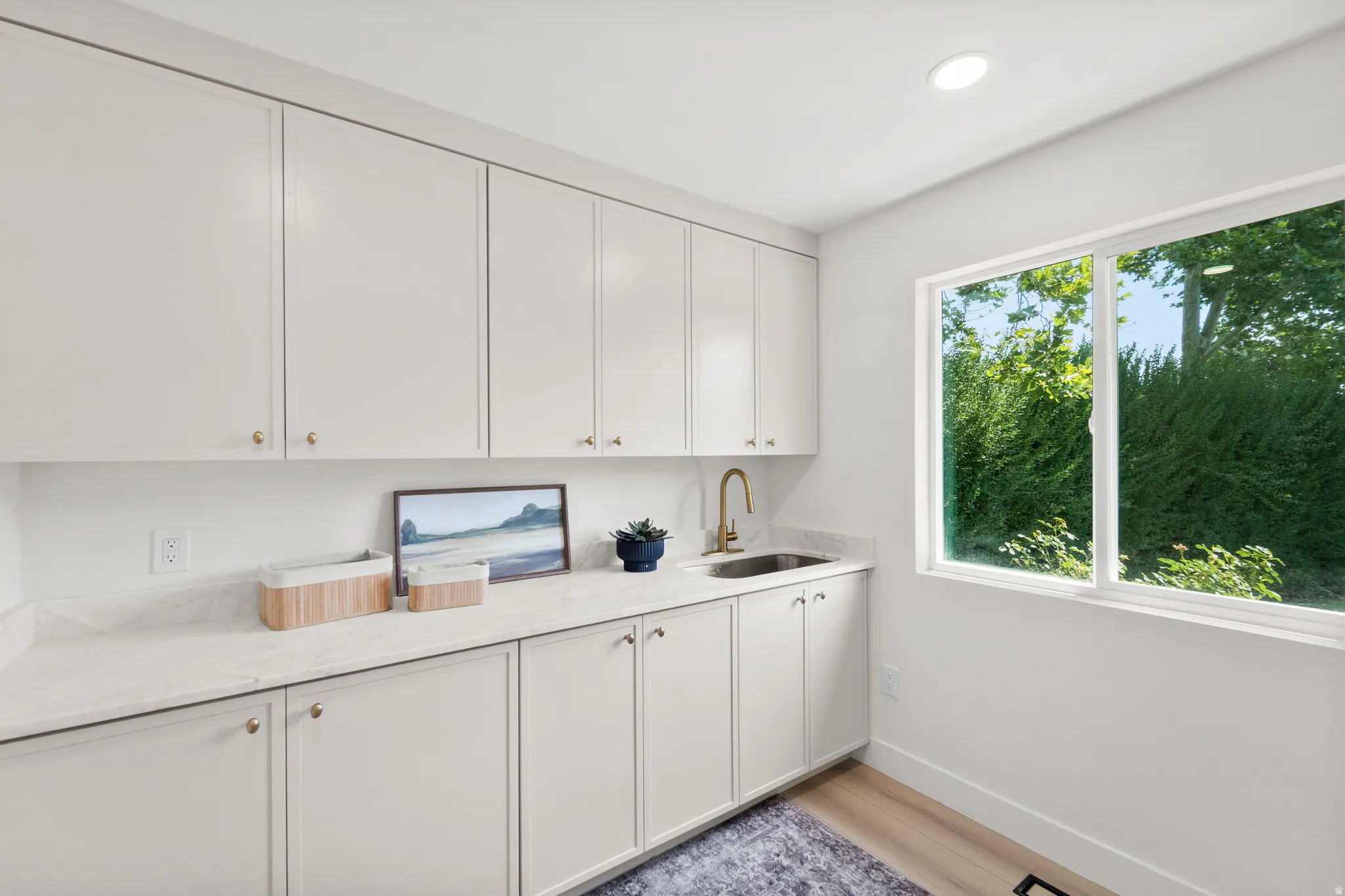 Extensive white cabinetry, light-toned countertops, integrated sink with gold-tone faucet, wood-finish flooring, and a large window
