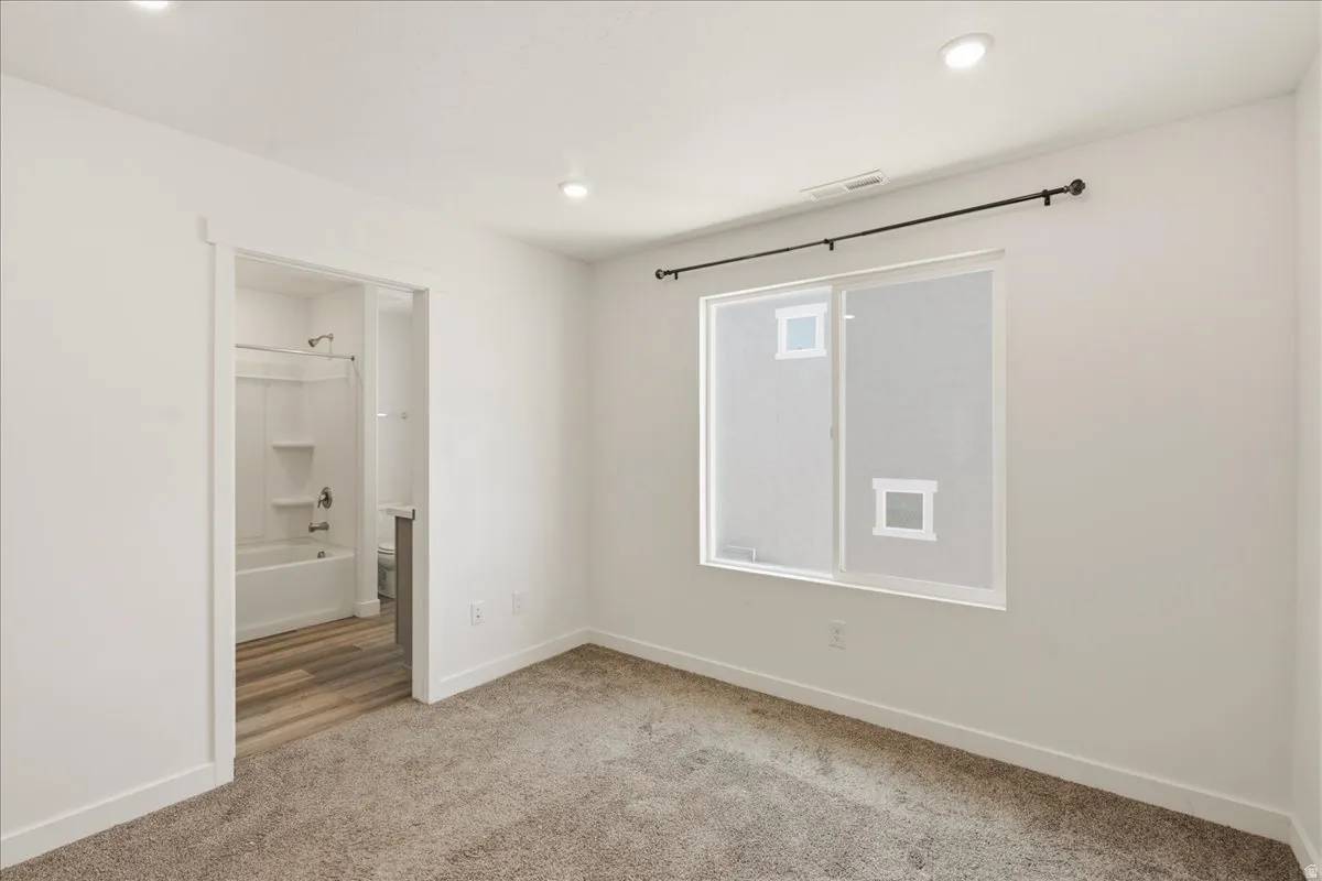 Carpeted room with recessed lighting and a sliding window, featuring a bathroom with wood-finish flooring and a bathtub
