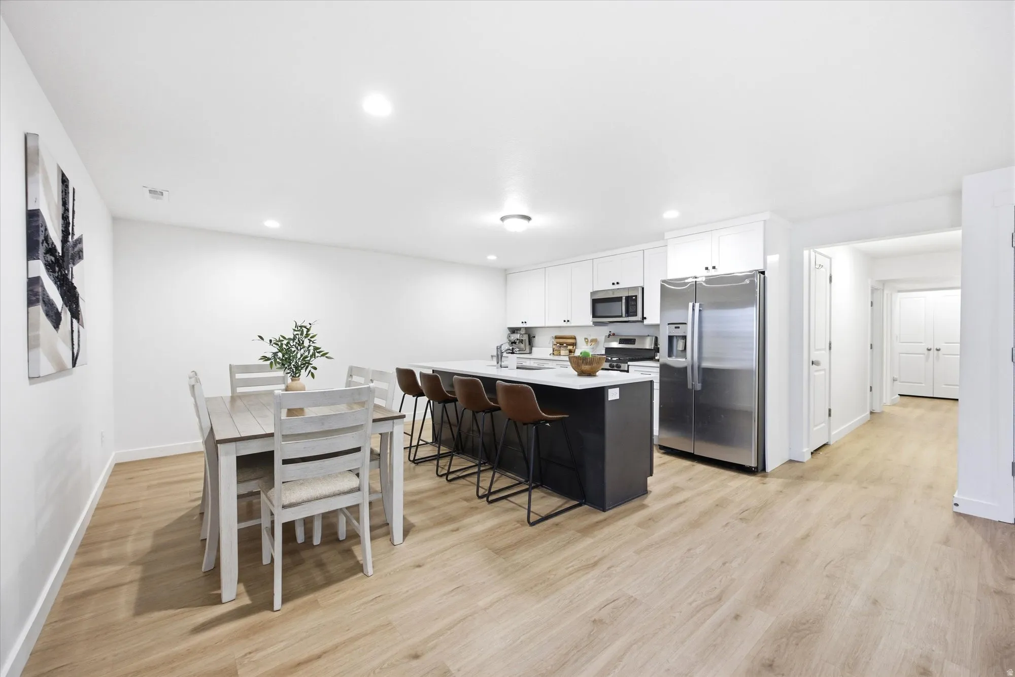 Open-concept living area featuring wood-finish flooring, recessed lighting, and a kitchen with stainless steel appliances, white cabinetry, and a central island with countertop seating