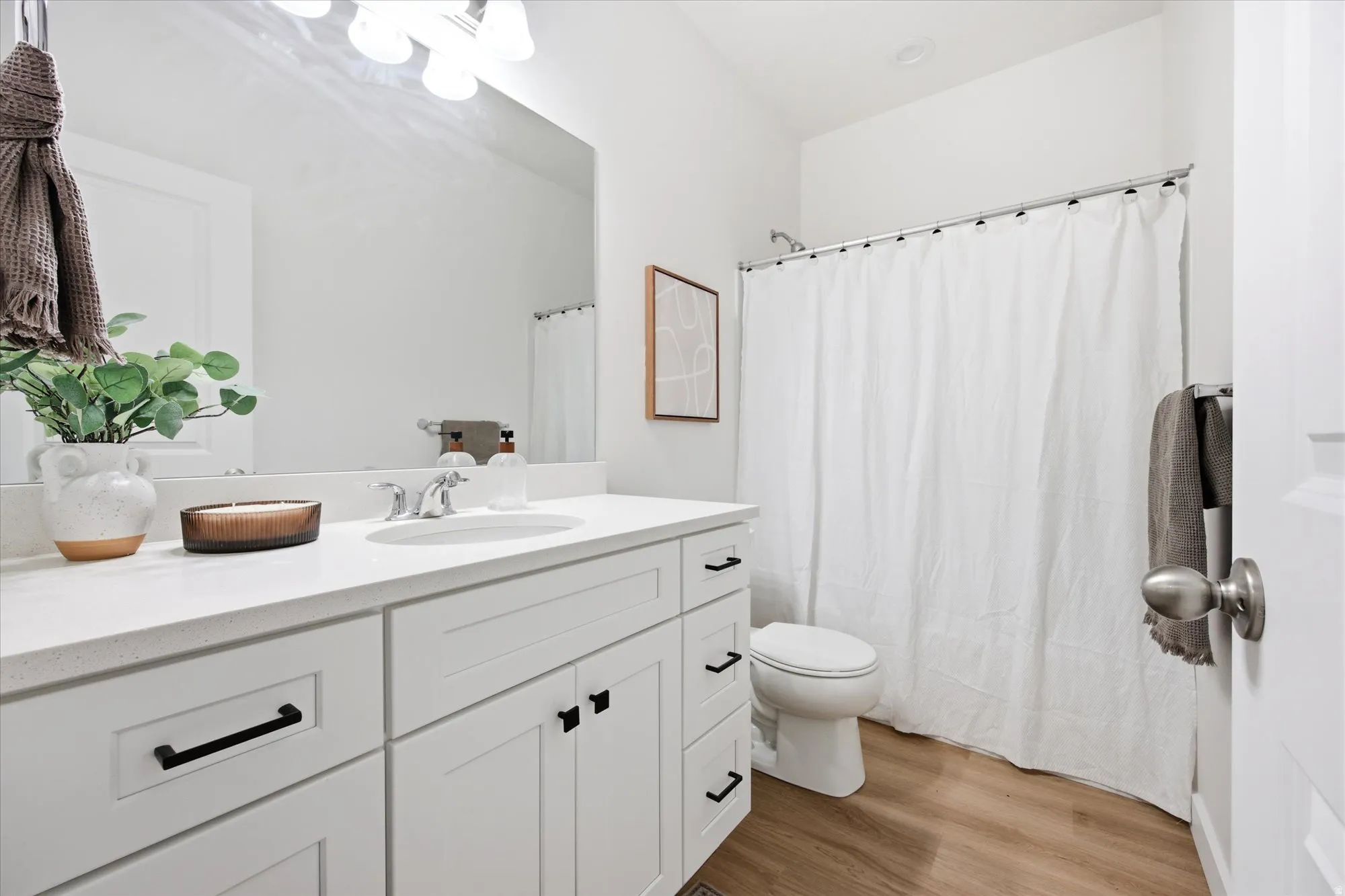 White vanity with black hardware, white countertop, undermount sink, overhead vanity lighting, and wood-finish flooring
