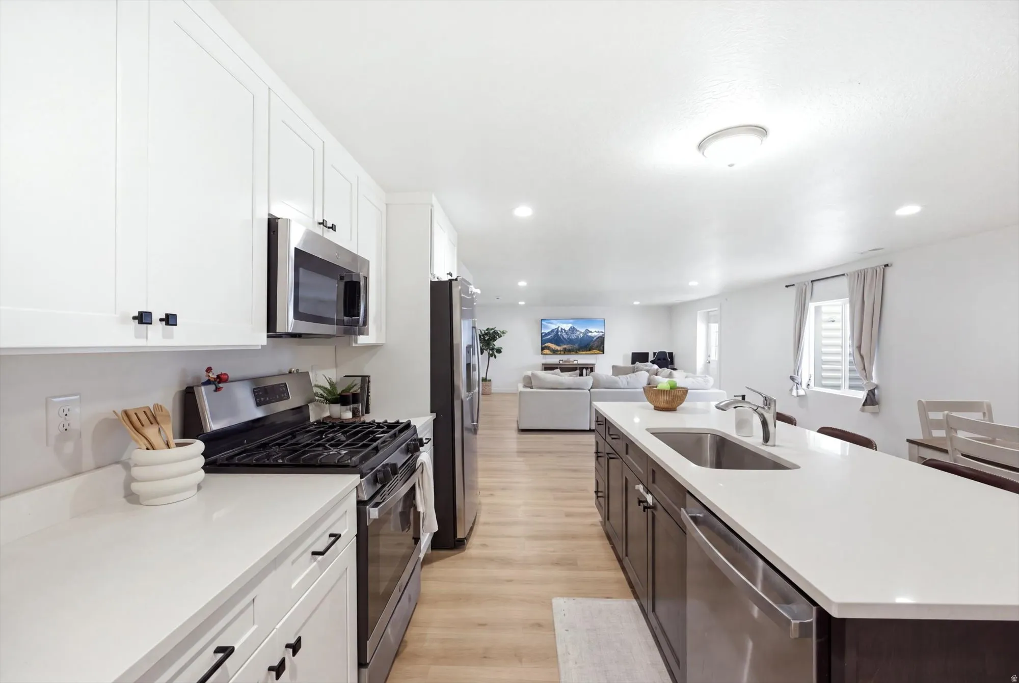 Kitchen featuring white shaker cabinetry, stainless steel appliances, light-toned countertops, a kitchen island with an integrated sink, and wood-finish flooring