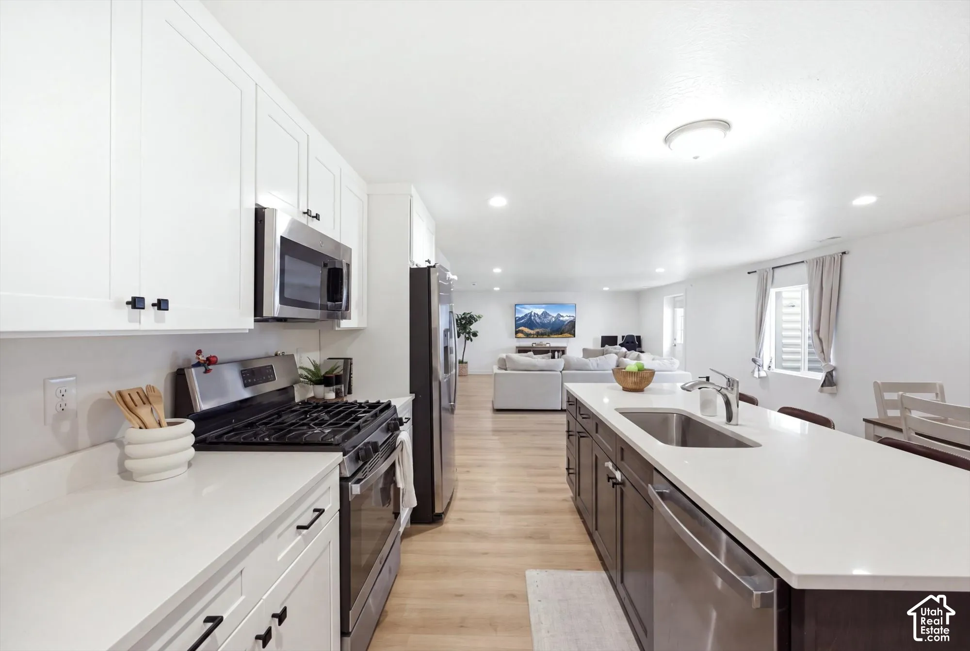 Kitchen featuring white shaker cabinetry, stainless steel appliances, light-toned countertops, a kitchen island with an integrated sink, and wood-finish flooring