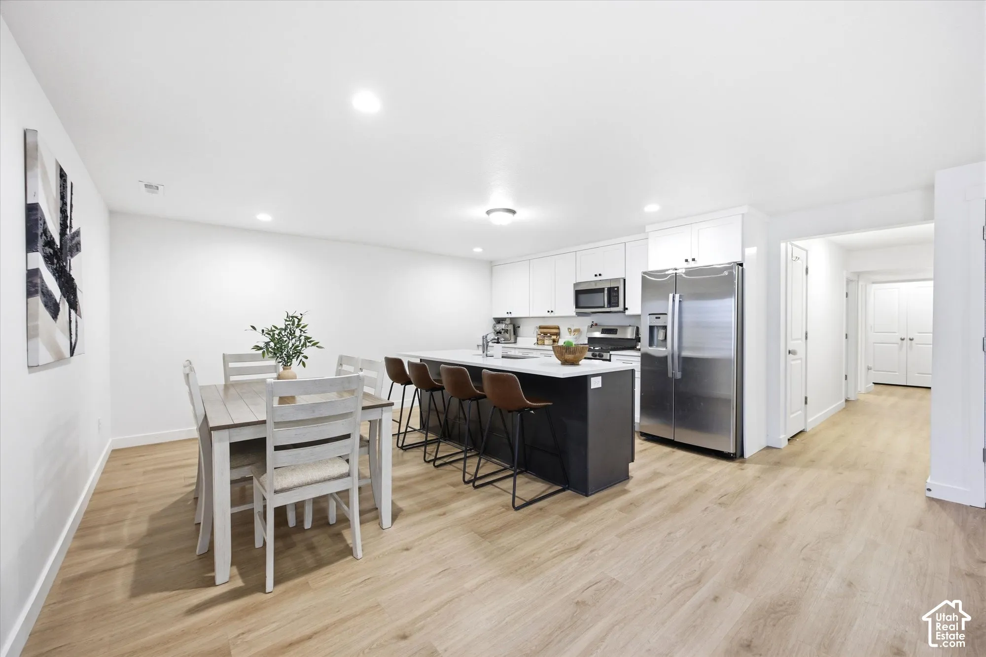 Open-concept living area featuring wood-finish flooring, recessed lighting, and a kitchen with stainless steel appliances, white cabinetry, and a central island with countertop seating