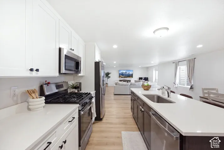 Kitchen featuring white shaker cabinetry, stainless steel appliances, light-toned countertops, a kitchen island with an integrated sink, and wood-finish flooring
