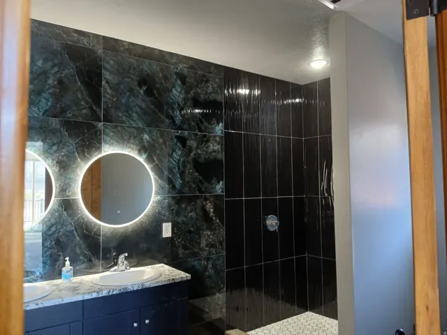Dual illuminated circular mirrors, dark stone-look wall tiling, double vanity with light-colored countertop, and a shower with dark rectangular wall tiling and white hexagonal floor tiling