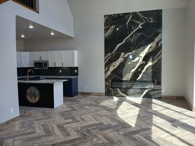 Herringbone wood-finish flooring, vaulted ceiling, built-in electric fireplace with a black and white veined tile surround, kitchen island with granite countertop, and white upper cabinetry
