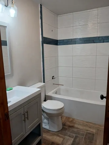 Bathroom featuring a white bathtub, wave-textured wall tiles with a decorative accent band, a vanity with a white countertop and integrated sink, a toilet, and wood-finish flooring