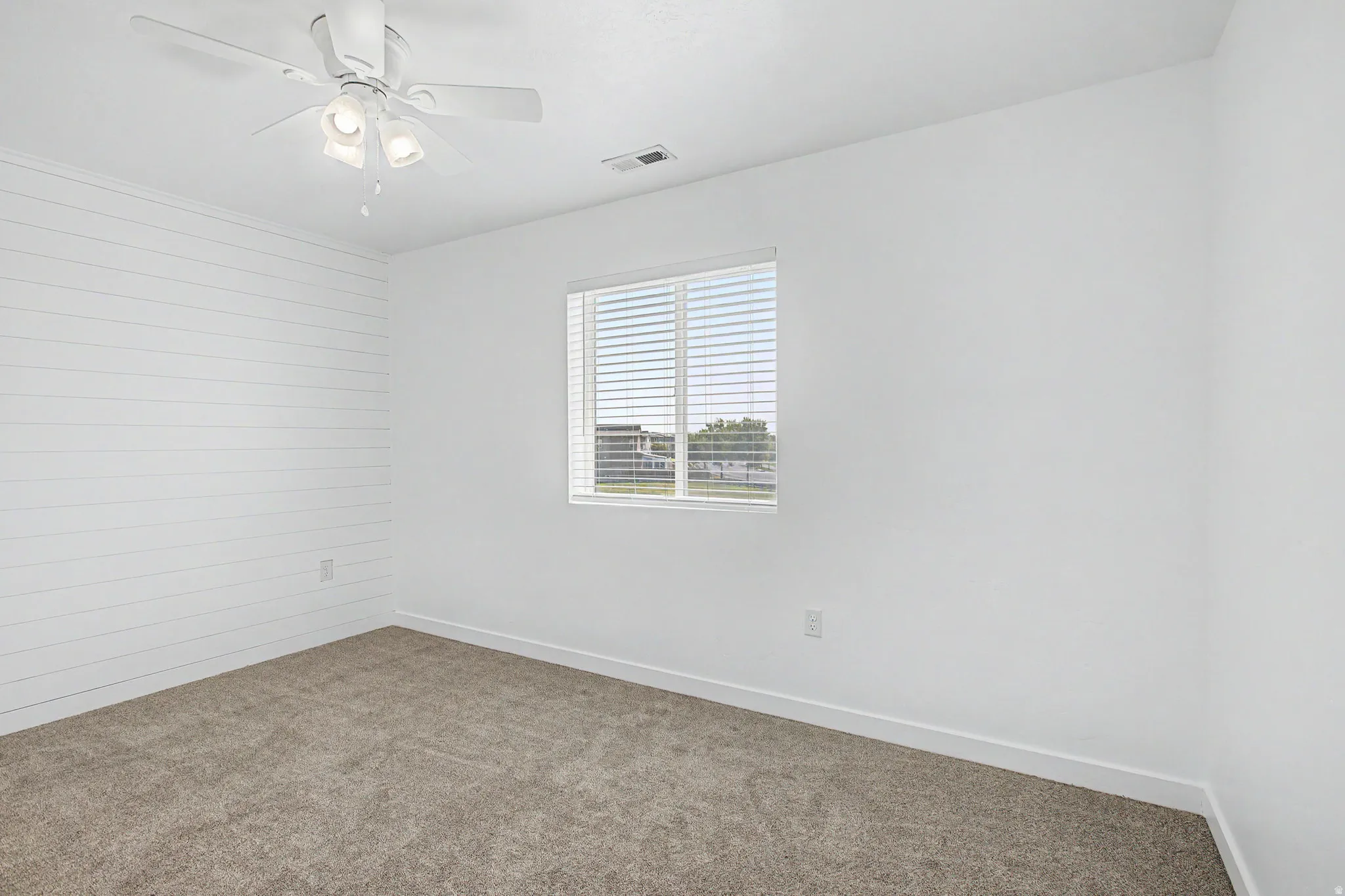Wall-to-wall carpeting with a bright window featuring blinds, a ceiling fan with integrated lighting, and a prominent shiplap accent wall