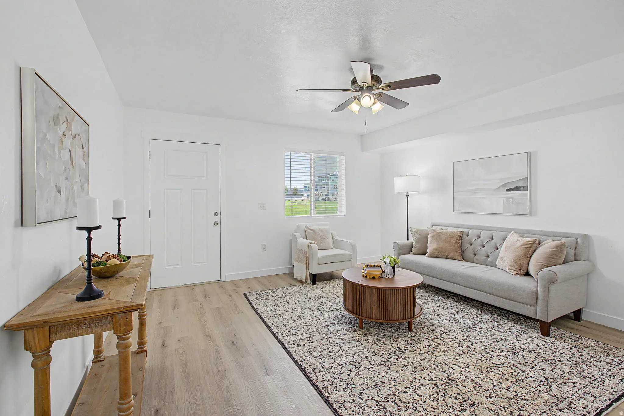 Spacious living area featuring light-toned wood-finish flooring, white walls, and a ceiling fan with integrated lighting