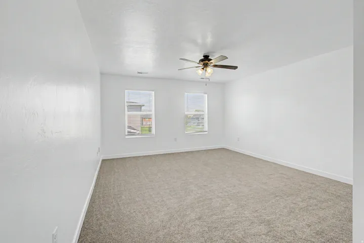 White walls, neutral carpet flooring, two windows with blinds, and a ceiling fan with integrated lighting