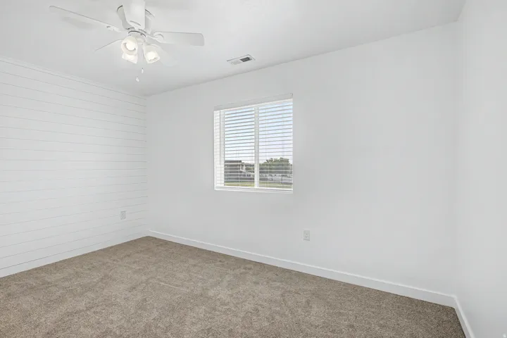 Wall-to-wall carpeting with a bright window featuring blinds, a ceiling fan with integrated lighting, and a prominent shiplap accent wall