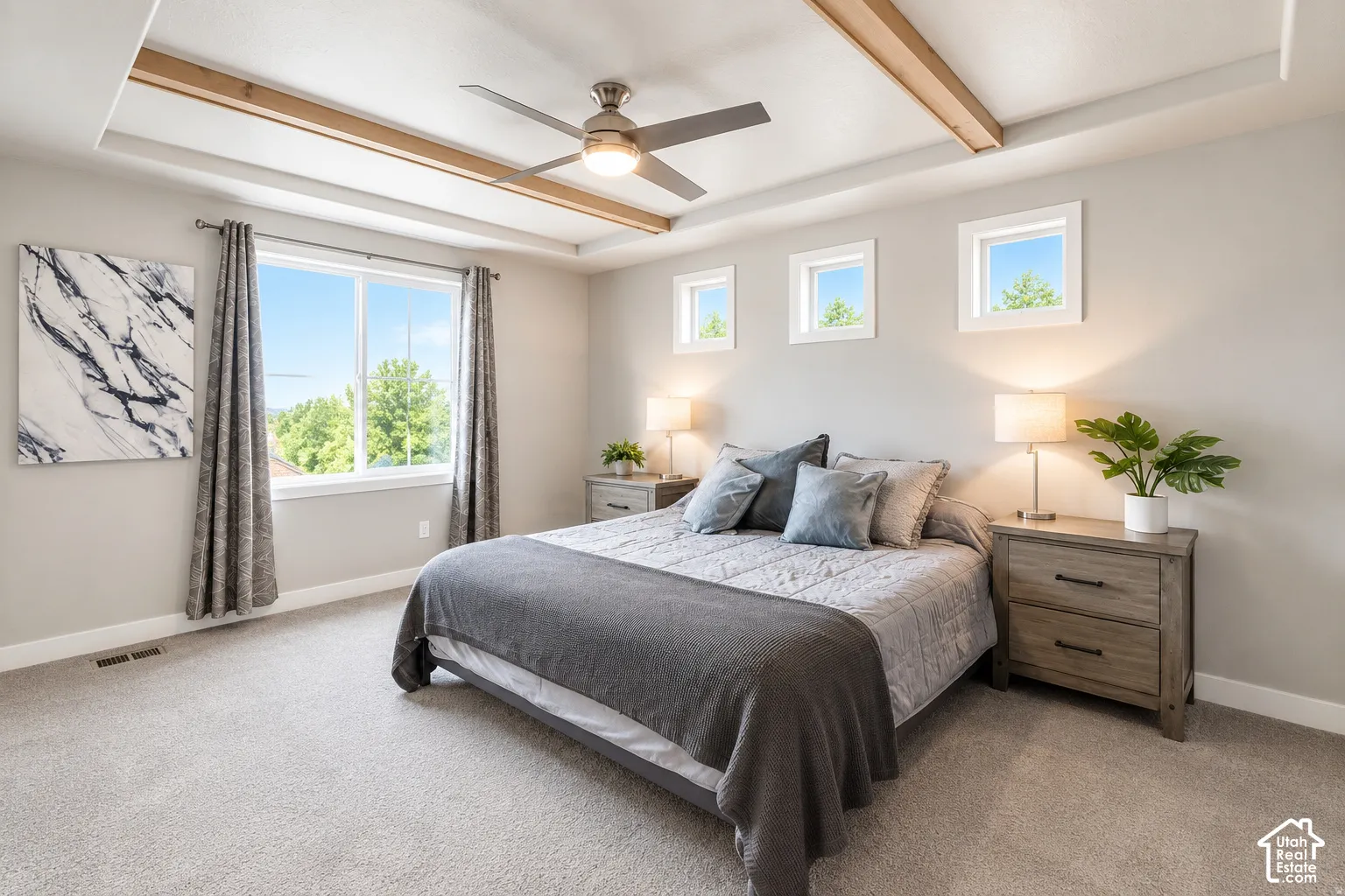 Primary bedroom tray ceiling with wood beam accents, ceiling fan with integrated lighting, multiple windows, light gray walls, and light gray carpeting