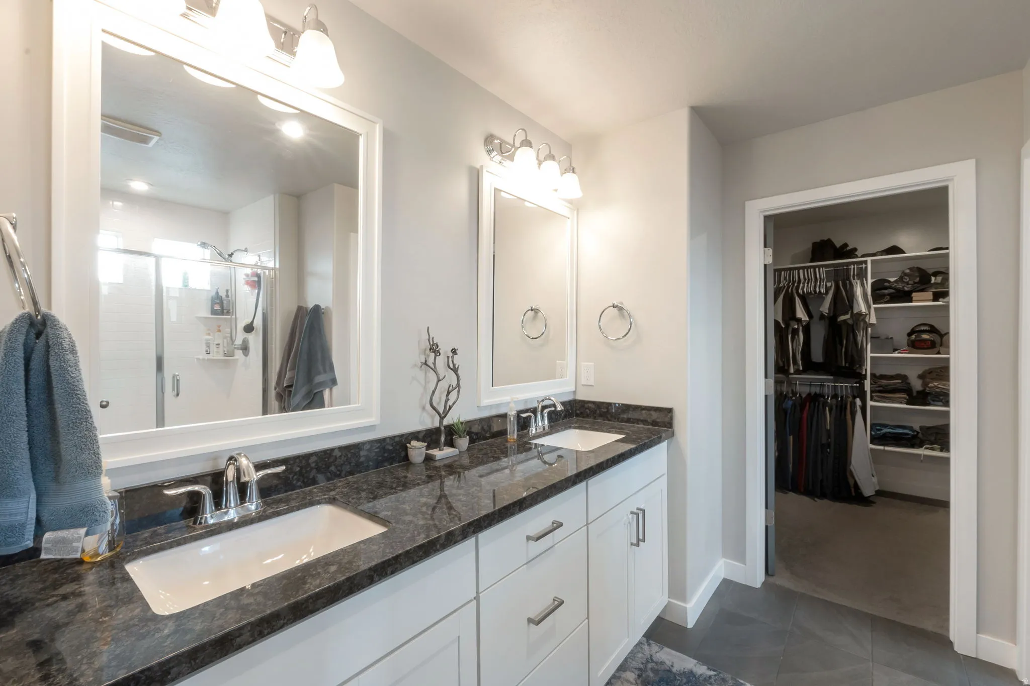 Spa-inspired primary bathroom featuring a large framed mirror with integrated lighting, dual-sink vanity with white cabinetry and brushed nickel hardware, dark stone countertops, and polished chrome fixtures
