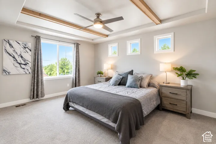 Primary bedroom tray ceiling with wood beam accents, ceiling fan with integrated lighting, multiple windows, light gray walls, and light gray carpeting
