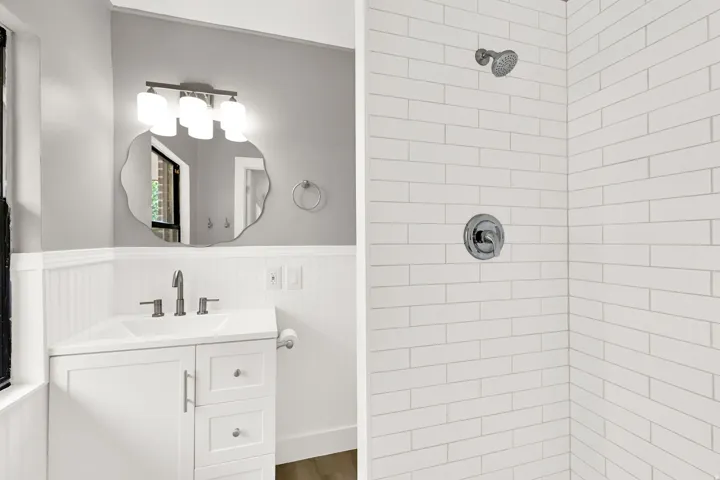White subway tile shower surround, chrome shower fixtures, white vanity with integrated sink, modern vanity light fixture, and decorative wall mirror