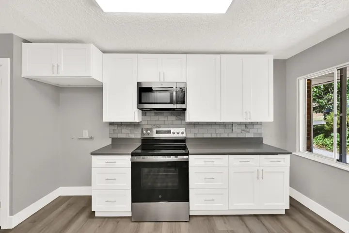 White shaker-style cabinetry, stainless steel oven with an integrated microwave, gray subway tile backsplash, dark countertops, and wood-finish flooring