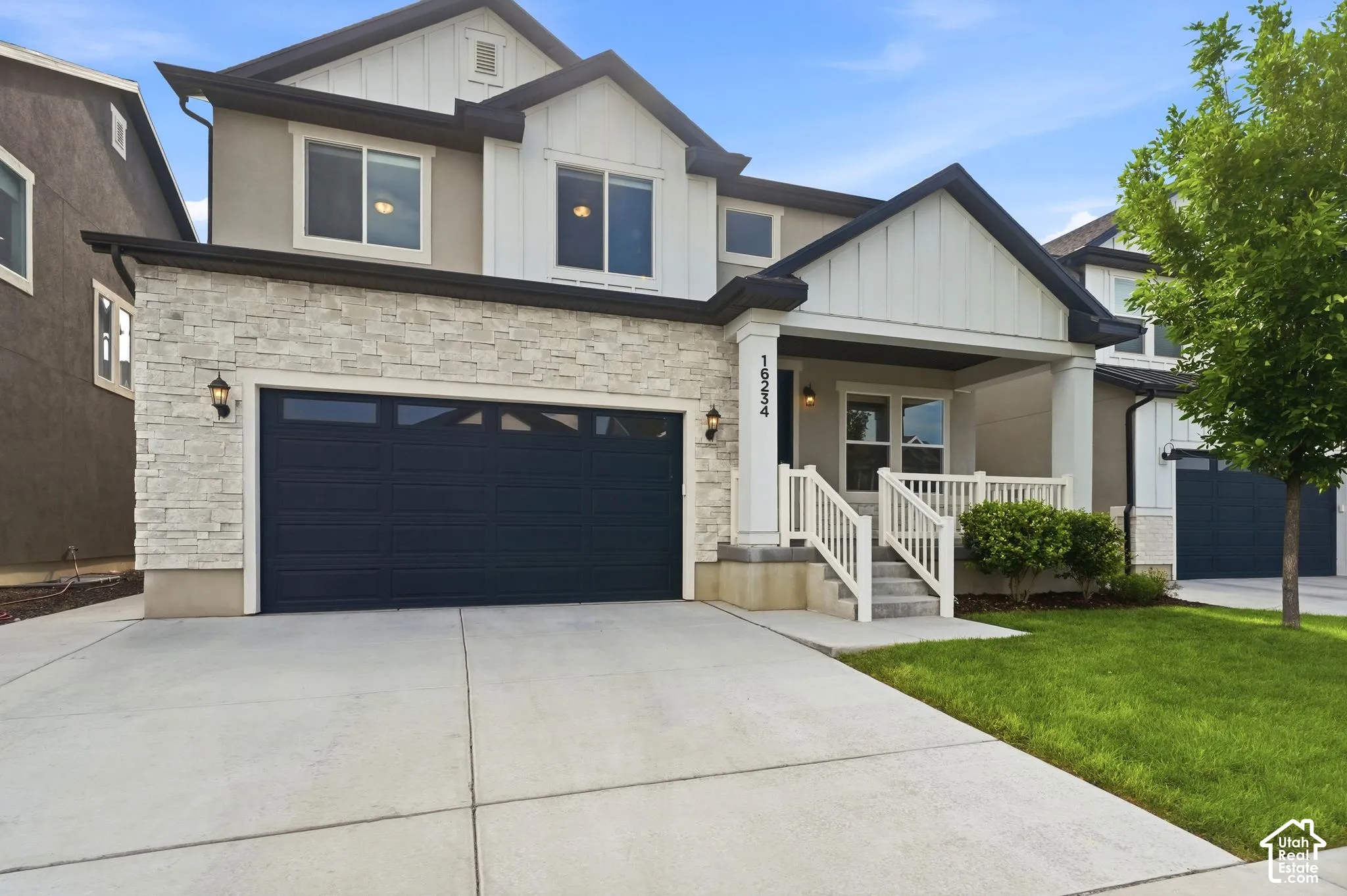 Contemporary two-story residence featuring a stone and stucco facade, a dark paneled garage door with upper window inserts, an inviting covered porch with white columns and railings, a concrete driveway, and a manicured lawn