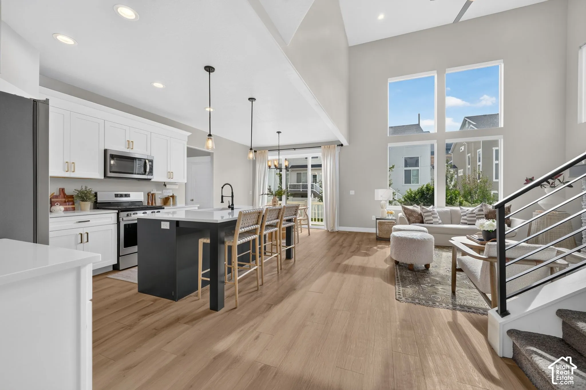 Open-concept living space with wood-finish flooring, a kitchen island with seating, a stainless steel appliance suite, a two-story ceiling, and a modern staircase with metal railings