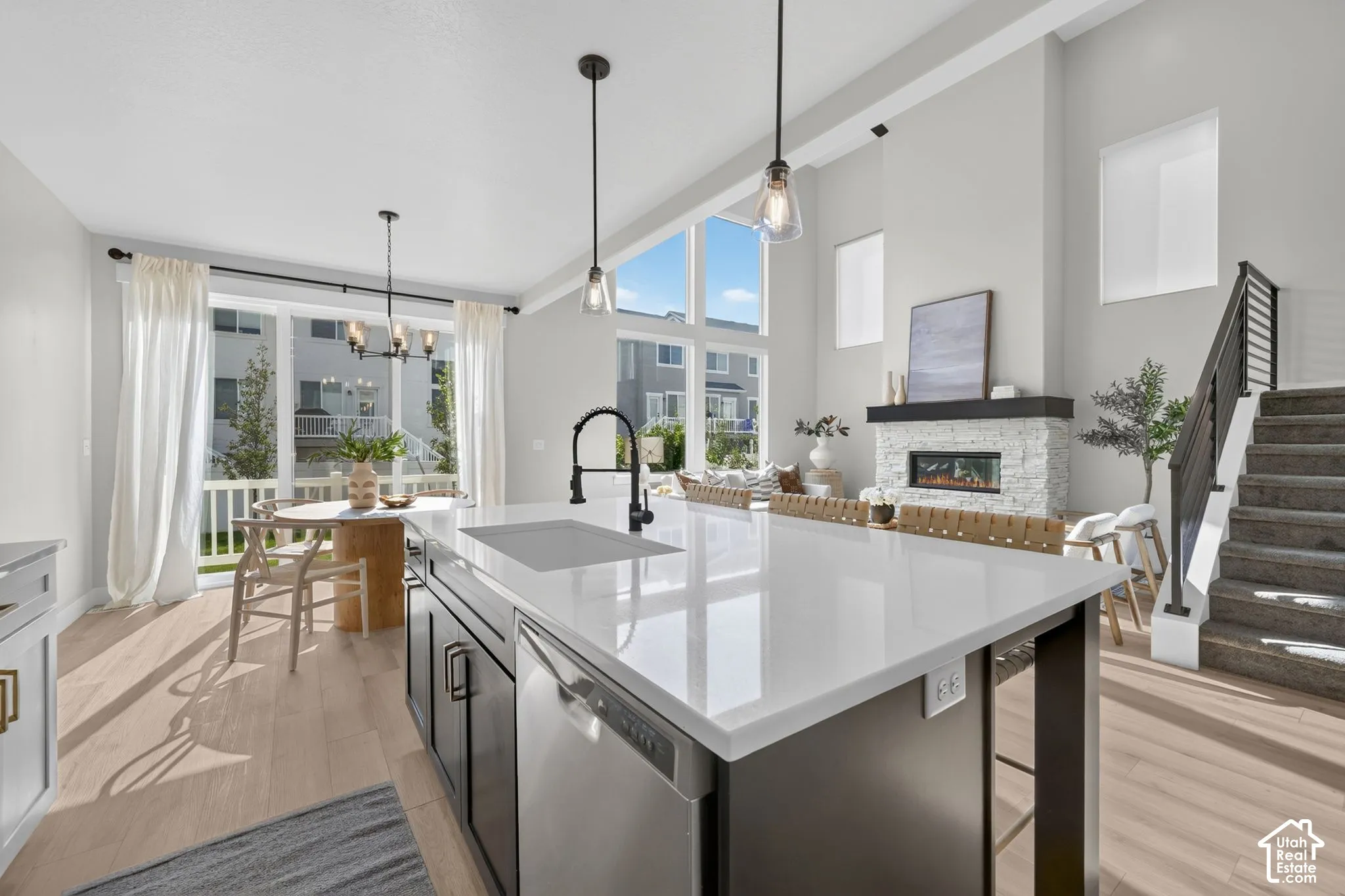White quartz island with integrated sink, stainless steel dishwasher, wood-finish flooring, two-story stone-clad fireplace, and multi-pane windows